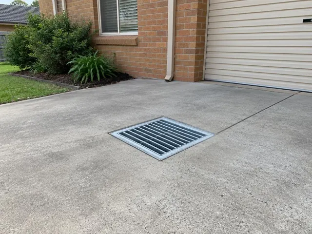 Clean outdoor driveway drain grate set in concrete beside a brick home and garage in Abbotsbury