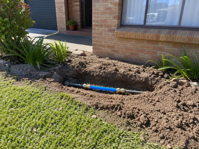 Open trench beside a brick house showing a repaired blue water pipe section in Abbotsbury
